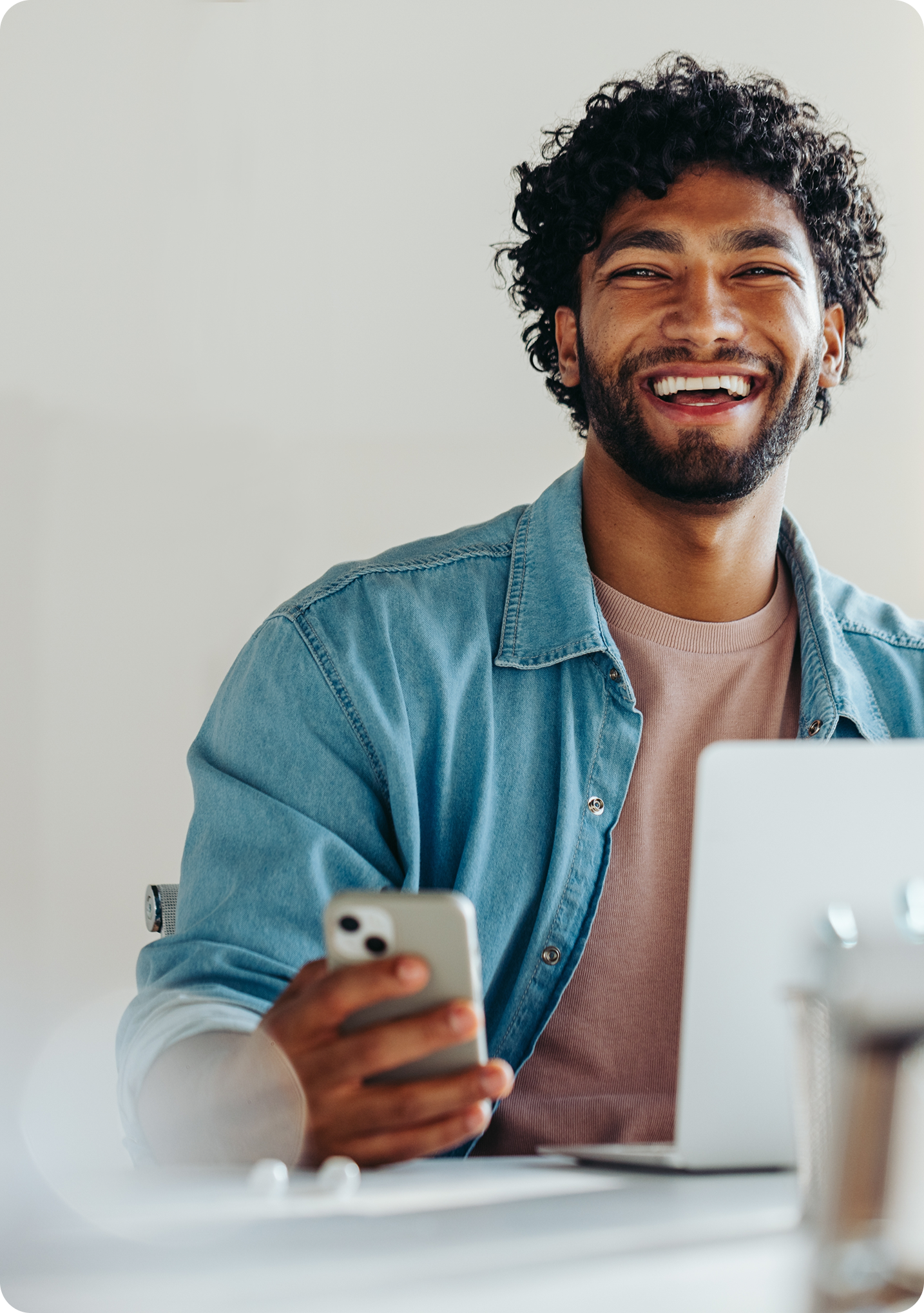 Image of a man in a teal shirt holding a mobile phone in his hand smiling towards the camera sat behind a computer.
